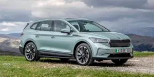 A green Skoda Enyaq electric SUV parked in a field with a mountain range in the background. The car has a modern and sleek design with black accents.