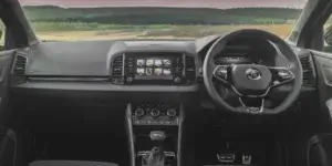 The interior of a Skoda Karoq SUV. The car features a black dashboard with a large central touchscreen display, a leather-wrapped steering wheel, and comfortable-looking front seats. The view outside the windshield shows a rural landscape with rolling hills and green fields.