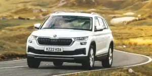 A white Skoda Karoq SUV driving down a winding road in a mountainous landscape. The car has a rugged exterior with black accents and roof rails.