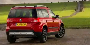 A red Skoda Yeti SUV driving down a winding road with a rural landscape in the background. The rear of the car is visible, showing the taillights, roof rails, and alloy wheels.