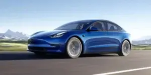 A blue Tesla Model 3 electric sedan driving on a highway with a scenic mountain landscape in the background. The car is in motion, and the background is blurred due to the speed.