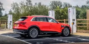 A red Audi Q8 e-tron electric SUV charging at an IONITY charging station. The car is plugged into one of the charging points, and the charging cable is visible. The background shows several other charging points and a fence. The license plate on the car is YN69 UYY.