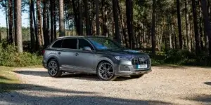 A gray Audi Q7 SUV parked in a forest. The car has a large size, a sporty design with a sloping roofline, and large alloy wheels. The license plate on the car is RMD 600.