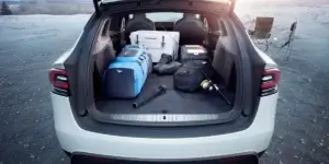 The open boot of a Tesla Model X electric SUV. The trunk is filled with camping gear, including a cooler, backpacks, hiking boots, and fishing rods. The trunk has a spacious interior and a flat load floor.