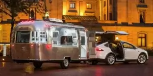 A white Tesla Model X electric SUV is parked next to a silver Airstream trailer. The trailer has its side door open, revealing a glimpse of the interior. The scene is set at night, with a building illuminated in the background.