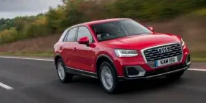 A red Audi Q2 SUV driving on a highway. The car has a sporty design with large alloy wheels and a prominent grille. The license plate on the car is KAM 150.