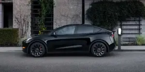 A black Tesla Model Y electric SUV parked in front of a modern building. The car is charging at a charging station and has black alloy wheels.