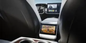 The rear seats of a Tesla Model X electric SUV. The seats are upholstered in white leather, and there is a center console with a built-in touchscreen display. The display shows a video game being played.