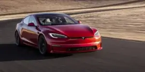 A red Tesla Model S Plaid electric sedan driving on a racetrack. The car is in motion, and the background is blurred due to the speed. The car has a sporty design with large alloy wheels and a low stance.