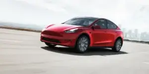 A red Tesla Model Y electric SUV driving on a highway with a cityscape in the background. The car is in motion, and the background is blurred due to the speed.