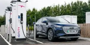 A blue Audi Q4 e-tron electric SUV charging at an IONITY charging station. The car is plugged into one of the charging points, and the charging cable is visible. The background shows several other charging points and a white building.