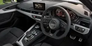 The interior of an Audi A4 car. The dashboard features a large touchscreen display, and the seats are upholstered in black leather. The steering wheel has multiple buttons for controls, and the center console includes a gear selector and other buttons.