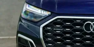 A close-up of the front of a blue Audi Q3 SUV. The car features distinctive LED headlights, a large honeycomb grille with the Audi logo, and the quattro badge indicating all-wheel drive.