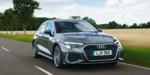 A gray Audi A3 Sportback car driving on a road with a field and trees in the background. The car has a sporty design with large alloy wheels and a prominent grille. The license plate on the car is LJV 360.