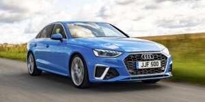 A blue Audi A4 sedan driving on a road with a field and trees in the background. The car has a sporty design with large alloy wheels and a prominent grille. The license plate on the car is JJF 500.