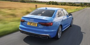 A blue Audi A4 sedan driving on a road with a field in the background. The car is in motion, and the rear of the car is visible, showing the taillights, rear bumper, and license plate. The license plate on the car is JJF 500.