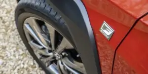 A close-up of a red car's front fender. The car has a black plastic fender flare and a chrome alloy wheel. The word "SPORT" is displayed on a badge on the fender flare.