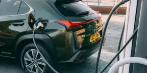 A green Lexus UX 300e electric SUV charging at an electric vehicle charging station. The charging cable is plugged into the car's charging port, and the car's rear is visible with the taillights and license plate. The license plate on the car is LZ72 UYD.