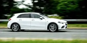 A white 2025 Mercedes-Benz A-Class hatchback car driving on a highway. The car has a sleek and modern design with alloy wheels. The background is blurred due to the car's speed.