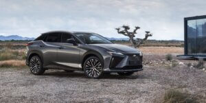 A gray Lexus RZ 450e electric SUV parked in a scenic landscape with mountains and a building in the background. The car has alloy wheels and a modern design.