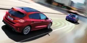 A red 2018 Ford Focus hatchback car driving on a busy city street. The car has a sporty design with alloy wheels and a rear spoiler. The car is using its radar sensors to detect other vehicles on the road, as shown by the green circles around the car.