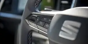 A close-up of a black leather steering wheel with silver accents. The steering wheel features buttons for cruise control, audio controls, and other functions. The SEAT logo is prominently displayed in the center.