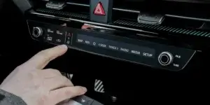 A close-up of a car's center console with a finger pressing a button labeled "AUTO." The console also features buttons for hazard lights, climate control, navigation, media playback, and radio tuning.