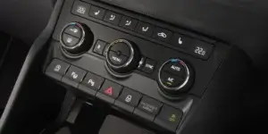 A close-up of the center console of a Skoda Karoq car. The console features a digital clock, climate control buttons, a hazard light button, a parking brake button, and a passenger airbag off button. The climate control buttons include a menu button, a sync button, and knobs for controlling temperature and fan speed.