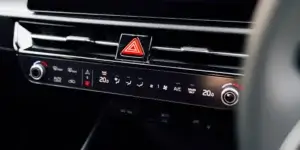 A close-up of the center console of a car. The console features a digital clock, a hazard light button, climate control buttons, and temperature knobs. The climate control buttons include a menu button, a sync button, and a button for controlling the passenger airbag. The temperature knobs are set to 20 degrees Celsius.