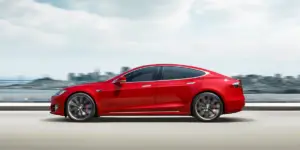 A red Tesla Model S electric car driving on a highway with a blurred cityscape and cloudy sky in the background.