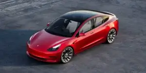 A red Tesla Model 3 electric sedan parked on a smooth gray surface, featuring a sleek aerodynamic design with black accents on the roof and wheels.