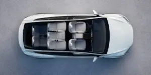 An aerial view of a white Tesla Model X electric SUV. The car is parked on a gray surface, and the interior is visible through the panoramic glass roof. The interior has three rows of seats with white leather upholstery.