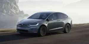 A grey Tesla Model X electric SUV driving on a scenic road with mountains and a misty lake in the background.