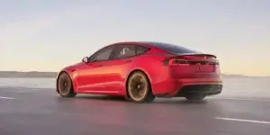 A red Tesla Model S Plaid electric sedan driving on a coastal road at sunset. The car is in motion, and the background is blurred due to the speed. The rear of the car is visible, showing the taillights, rear bumper, and license plate. The car has a sporty design with large alloy wheels and a rear spoiler.