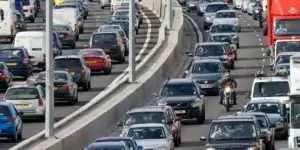 A busy highway with heavy traffic congestion. Cars, vans, and trucks are all lined up, bumper to bumper, and there is a motorcycle weaving through the traffic. The background shows a cityscape with tall buildings.