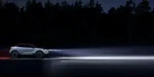 A silver Opel Mokka crossover SUV driving on a dark road at night. The car's headlights are on, illuminating the road ahead. The car is surrounded by trees and a starry sky.