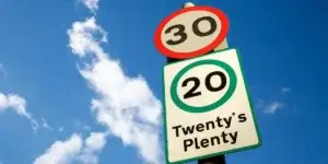 A road sign with a 30 mph speed limit sign above a 20 mph speed limit sign. The 20 mph sign also has the text "Twenty's Plenty" below it. The background is a blue sky with white clouds.