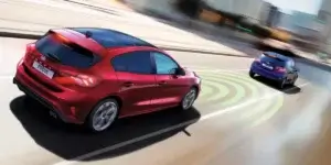 A red Ford Focus hatchback car driving on a busy city street. The car is using its radar sensors to detect other vehicles on the road, as shown by the green circles around the car. The background of the image is blurred due to the car's speed.