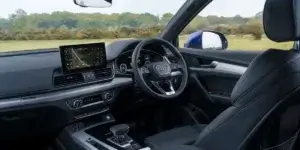 The interior of an Audi Q5. The car has a black leather interior, a large center screen with a navigation map displayed, a three-spoke steering wheel with the Audi logo, and a gear selector. The car appears to be parked in a scenic countryside location.