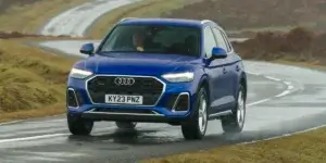 A blue Audi Q5 driving on a winding road in the countryside. The car is facing towards the viewer and has a license plate that reads "KY23 PNZ."