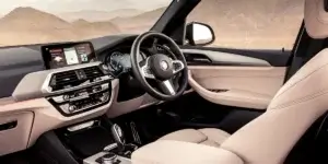 The interior of a BMW X3 SUV. The car features a beige leather interior, a large center screen, a three-spoke steering wheel with the BMW logo, and a gear selector. The car appears to be parked in a desert landscape.