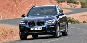 A blue BMW X3 driving on a winding road in a mountainous landscape. The car is facing towards the viewer and has a license plate that reads "YJ67 ZSG."