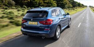 A blue BMW X3 driving on a winding road in the countryside. The car is facing away from the viewer and has a license plate that reads "YG20 DZK."