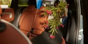 The backseat of a MINI Cooper Countryman car. The rear seats are folded down, and a large bouquet of pink flowers is placed in the center of the seat. A person is standing behind the car, reaching into the trunk to retrieve something. The car has a brown leather interior.