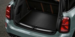 The open trunk of a MINI Cooper Countryman car. The trunk is empty and has a black rubber mat with a diamond pattern. The rear taillights are visible on either side of the trunk opening. The tailgate is open and propped up.