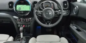 The interior of a MINI Cooper Countryman car. The dashboard features a large touchscreen display with a navigation map, a digital instrument cluster, and a leather-wrapped steering wheel. The seats are upholstered in a combination of black and white leather, and the center console has a gear selector and other buttons. The MINI logo is visible on the steering wheel.