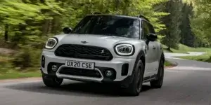 A white MINI Cooper Countryman driving on a winding road with trees on both sides. The car has black alloy wheels, a black roof, and a sporty design. The license plate on the car is OX20 CSE.