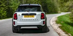 A white MINI Cooper Countryman SE electric SUV driving on a winding road with trees on both sides. The car has a sporty design with black accents and alloy wheels. The license plate on the car is OX20 CSE.