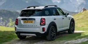 A white MINI Cooper Countryman driving on a winding road with a scenic mountain backdrop. The car has black alloy wheels, a black roof, and a sporty design. The license plate on the car is OX20 CSE.