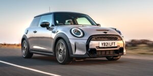 A silver MINI Cooper Electric car driving on a road with a scenic landscape in the background. The car has a modern design with black accents and alloy wheels. The license plate on the car is YB21 LFT.